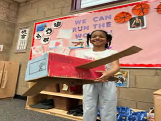 A young girl is holding a cardboard airplane in front of a bulletin board.
