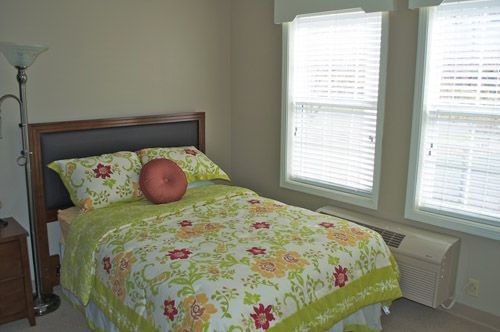 A bedroom with a bed , nightstand , lamp and two windows.