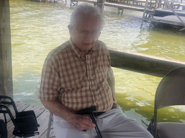 An elderly man is sitting on a chair next to a body of water.