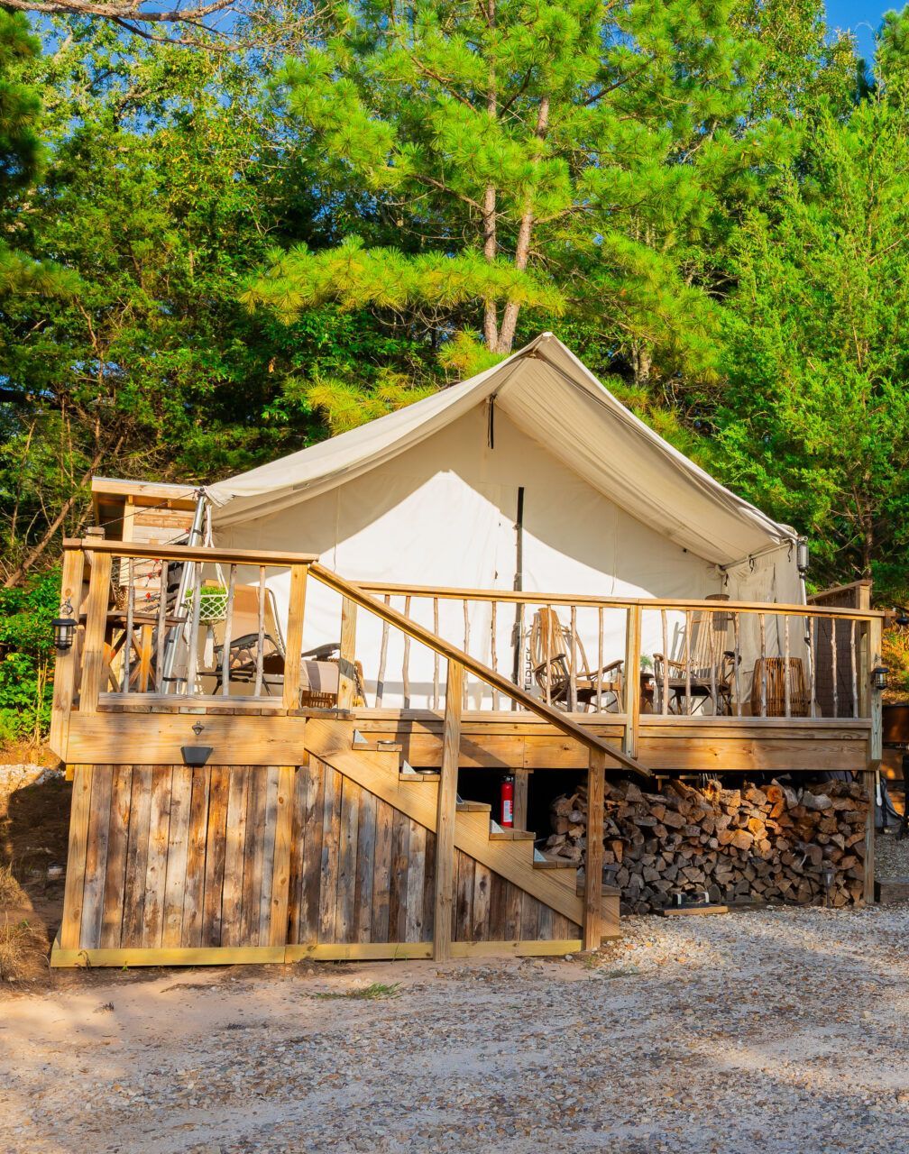 Canvas glamping tent with wooden deck and firewood, surrounded by green trees in a sunny forest setting