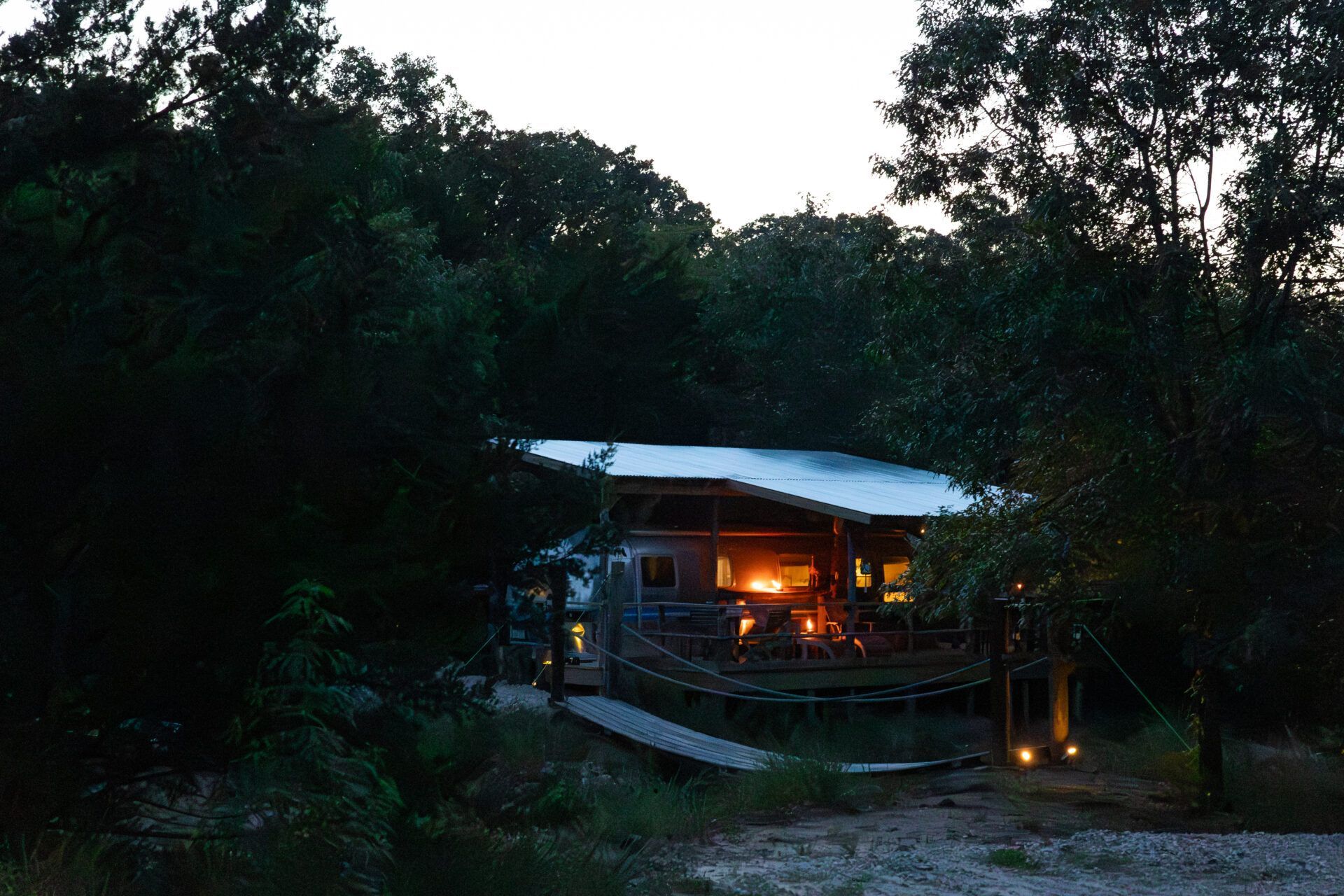 Small lit cabin in a dark forest at dusk, with warm lights glowing through the windows.