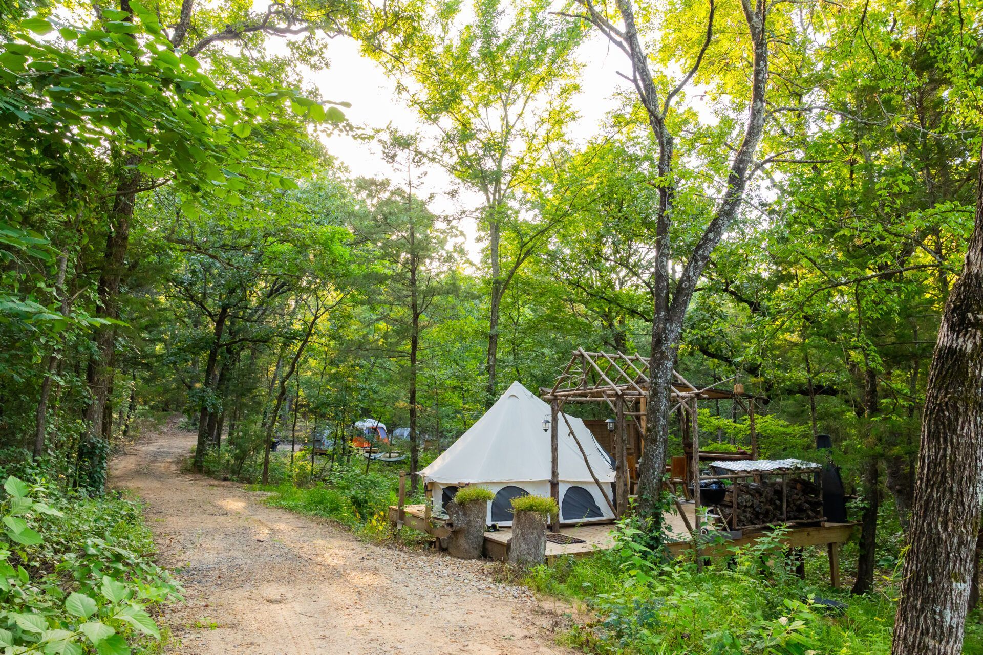 White tent in a wooded campsite beside a dirt path and parked vehicle