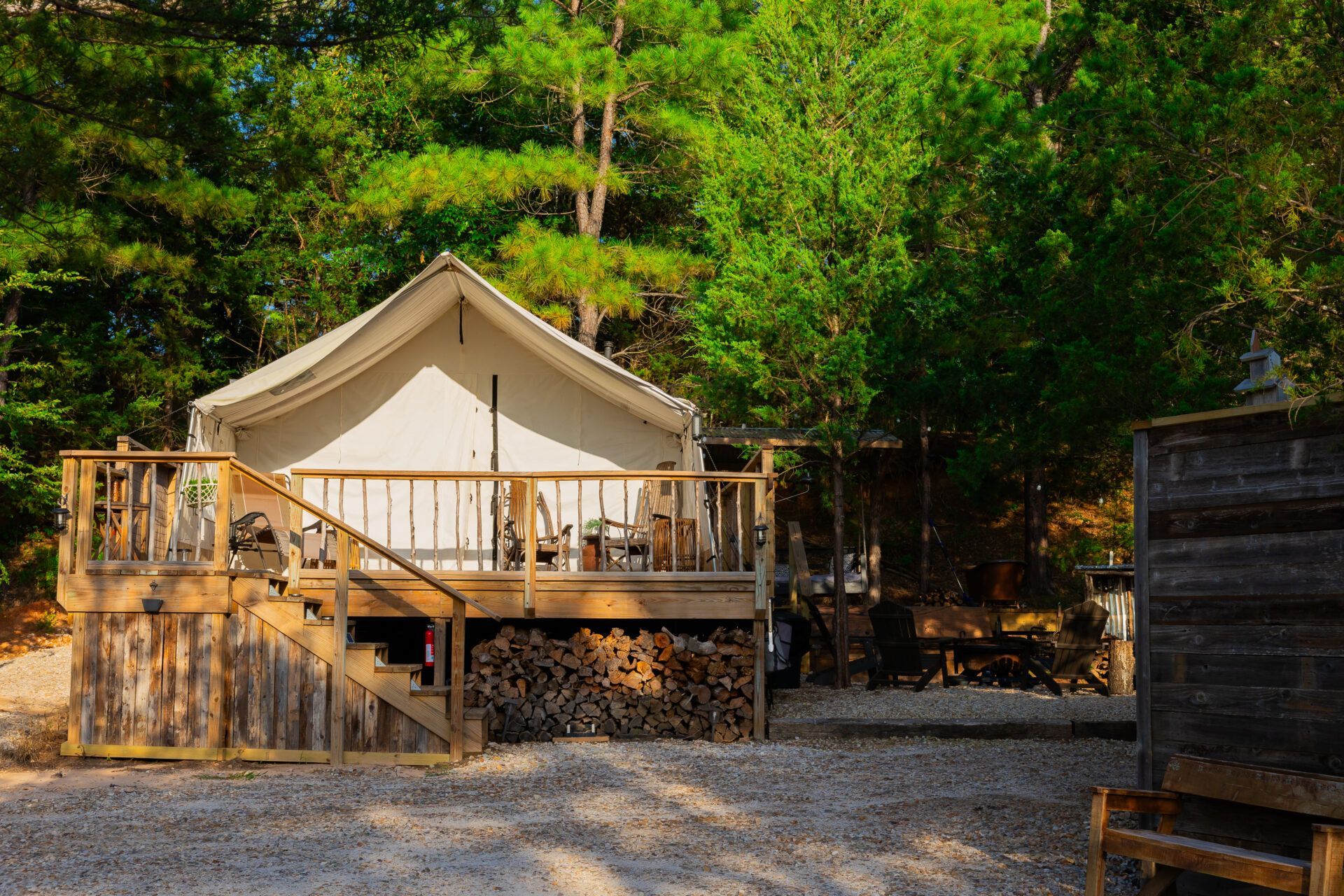 White canvas tent cabin on a wooden deck in a forest clearing, surrounded by trees and gravel.