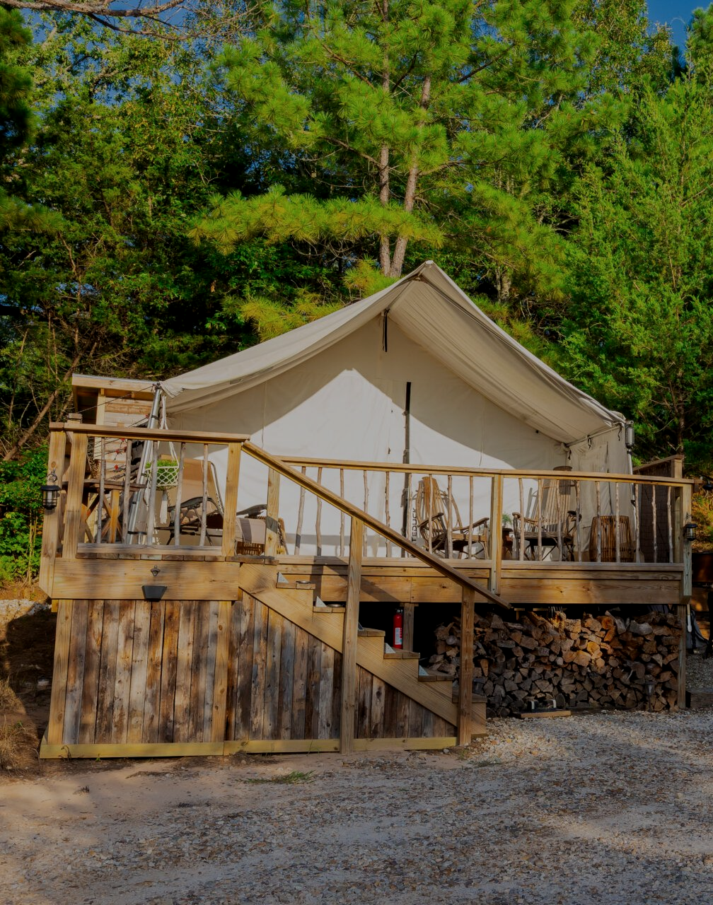 Canvas glamping tent on a wooden deck in a wooded campsite, with outdoor seating and firewood beneath.
