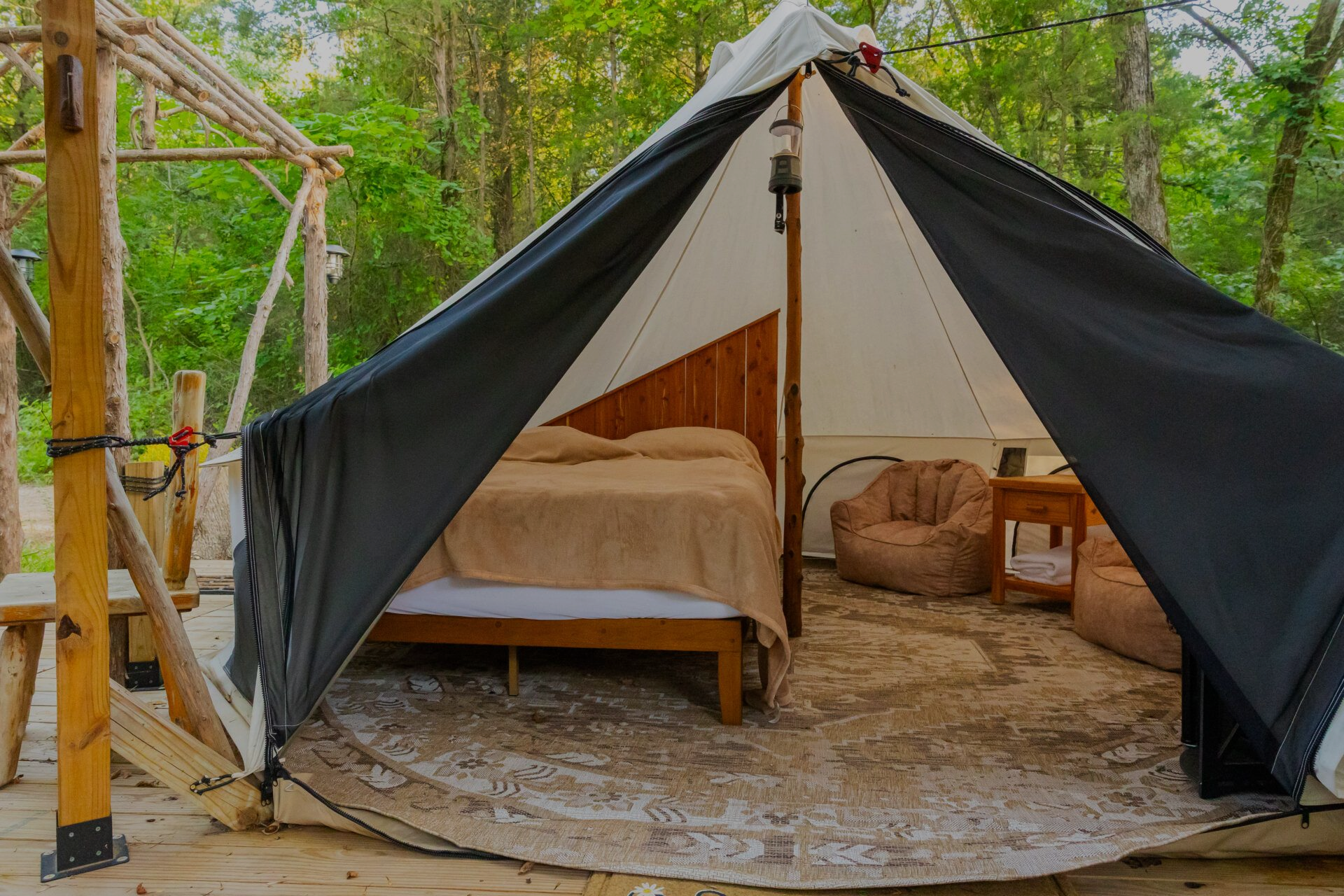 Tent-style glamping tent with wooden bed, beige bedding, and round rug in a lush outdoor setting
