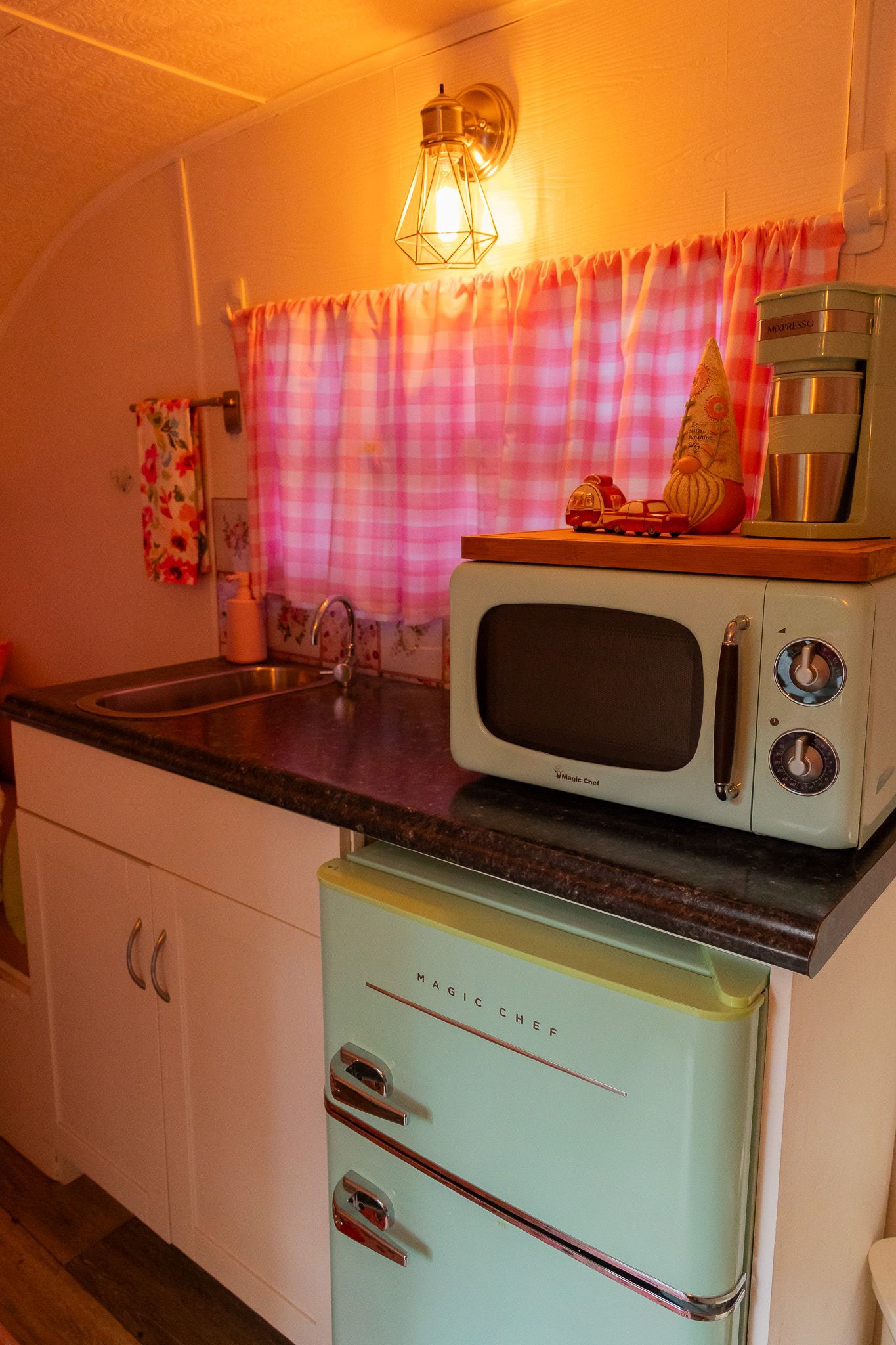 Small retro kitchen with a microwave on a countertop, a mini fridge below, and a pink-curtained window lit warmly.