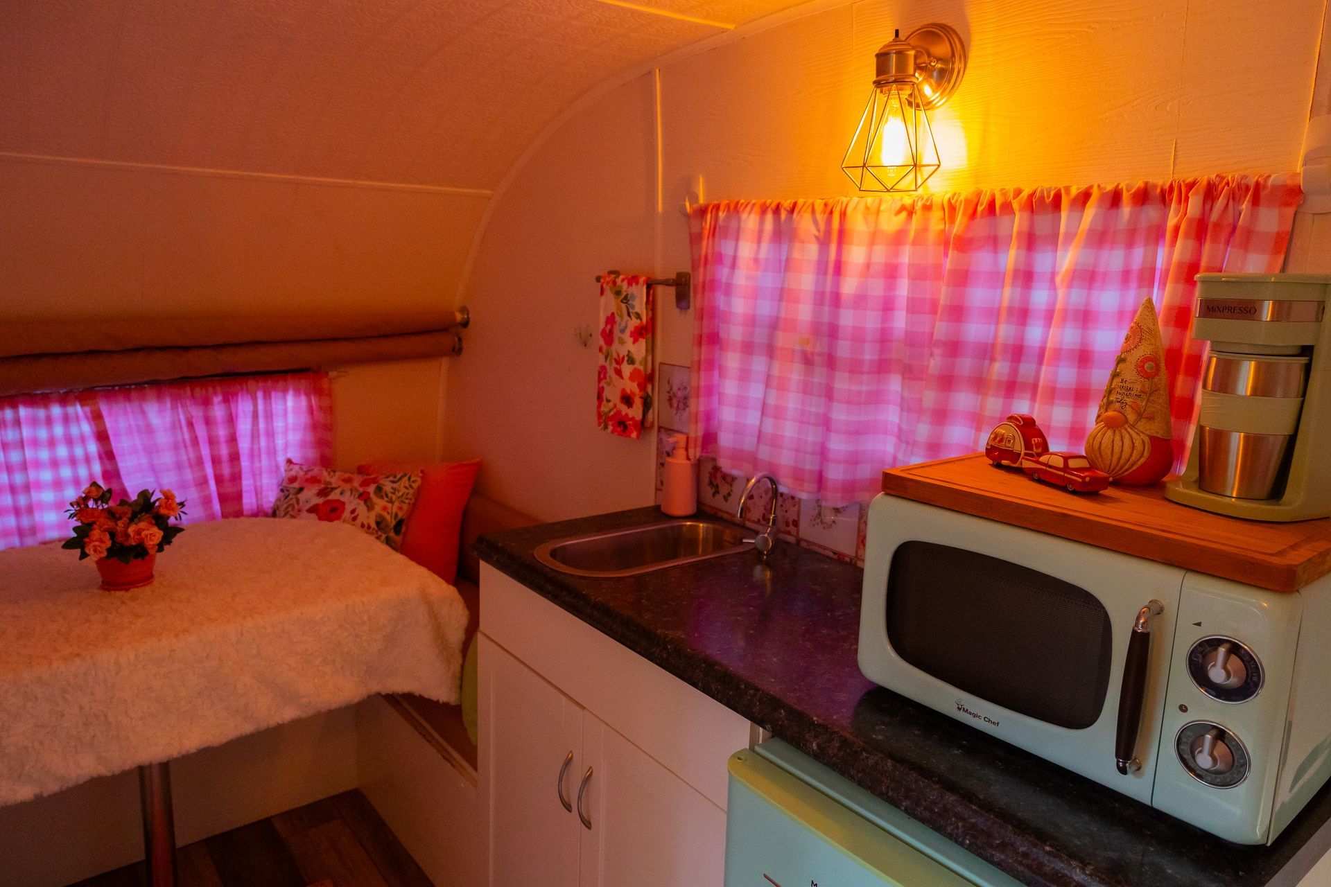 Small cozy kitchen nook with pink curtains, a microwave, sink, and table under warm lighting