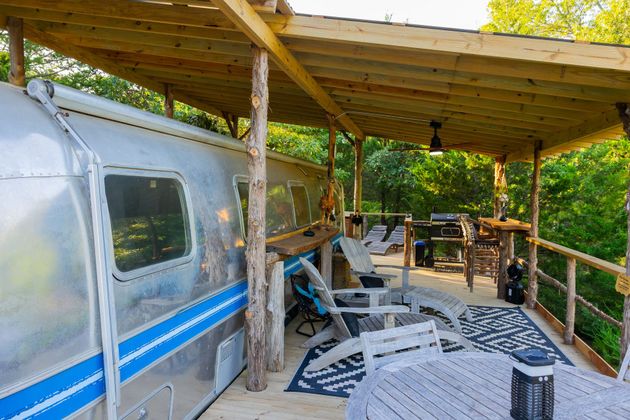 Covered outdoor deck beside a silver camper trailer with white chairs and wooden railing in a green forest setting