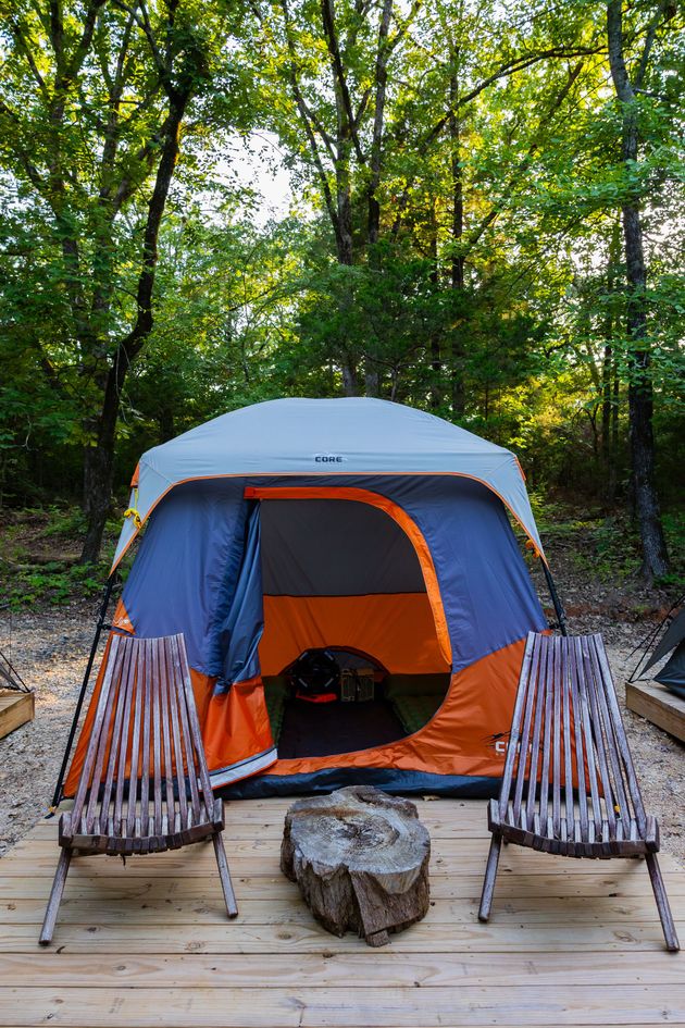 Tent on a wooden deck in a wooded campsite, flanked by two lounge chairs and a rock fire pit.