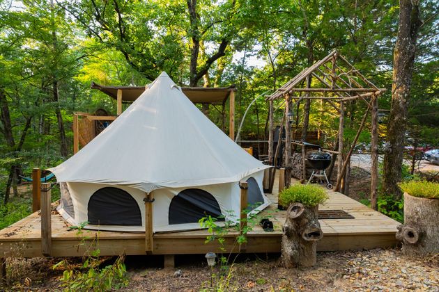 White canvas tent on a wooden platform in a wooded campsite, with another shelter and trees behind it
