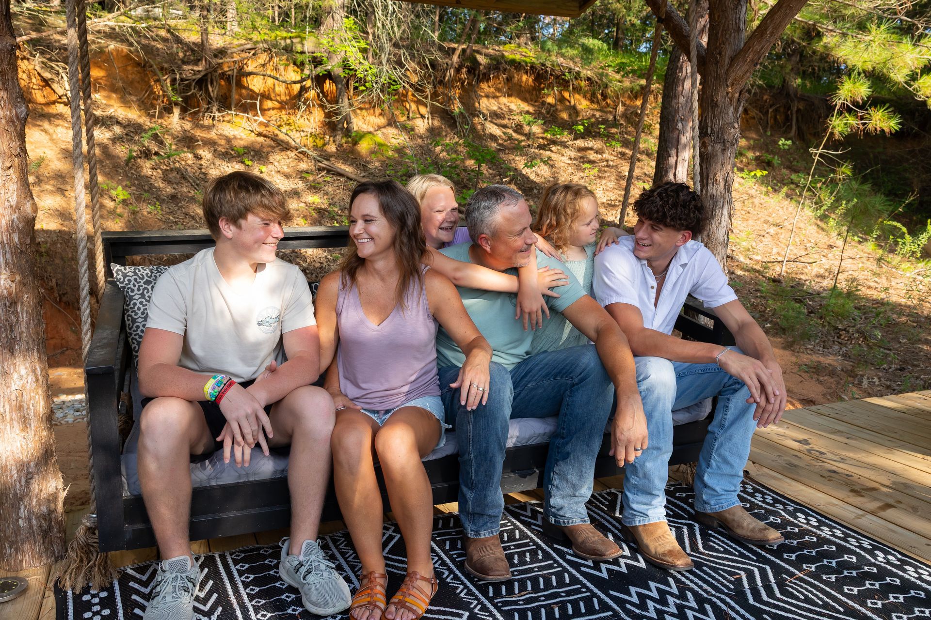 Four people sitting on an outdoor bench and rug, smiling in a sunny wooded area.