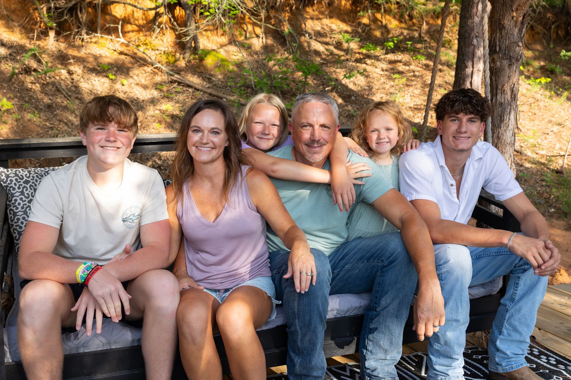 Six people sitting close together on a couch outdoors, smiling in a sunlit wooded area.