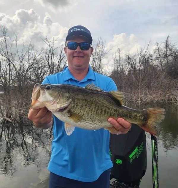 Man holding a large bass on a boat, wearing a blue shirt and cap on a calm lake