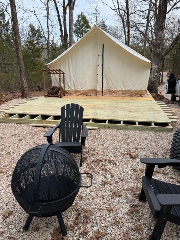 Outdoor campsite with cream-colored tent, wooden platform, and black chairs around a fire pit.