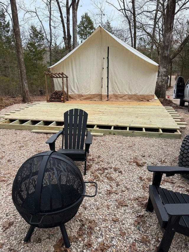 Outdoor campsite with cream-colored tent, wooden platform, and black chairs around a fire pit.