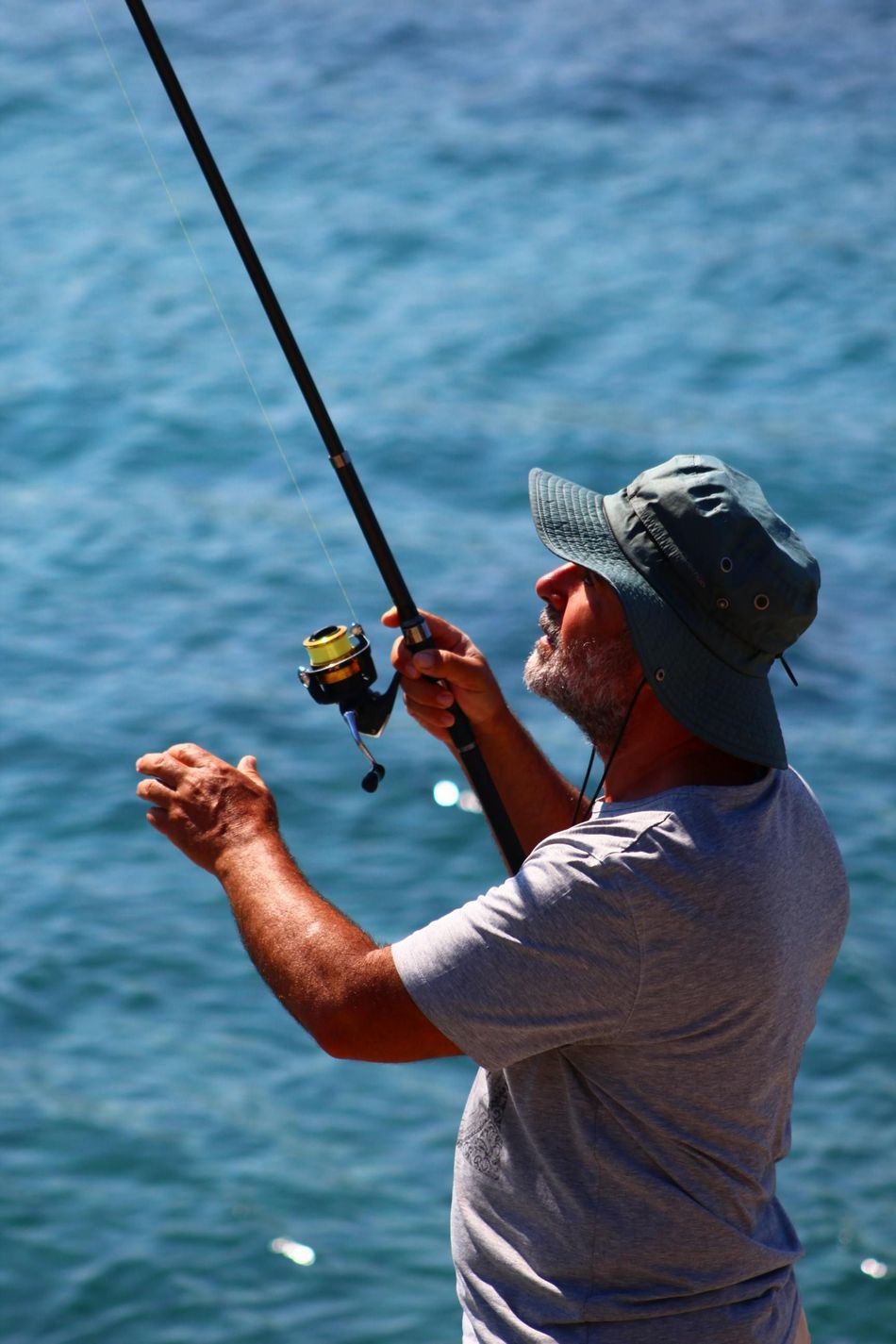 Man fishing from a boat, holding a rod over blue water and reeling in a catch
