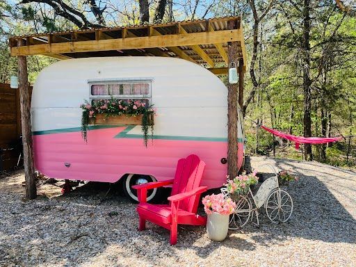 Pink and white tiny home with porch, red chairs, and flower pots in a wooded setting