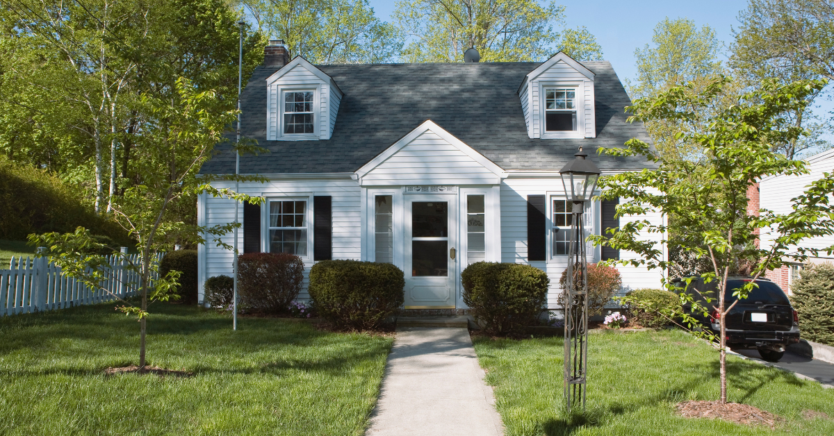 White Cape Cod-style house with a gray roof and dormer windows, surrounded by green trees and shrubs.