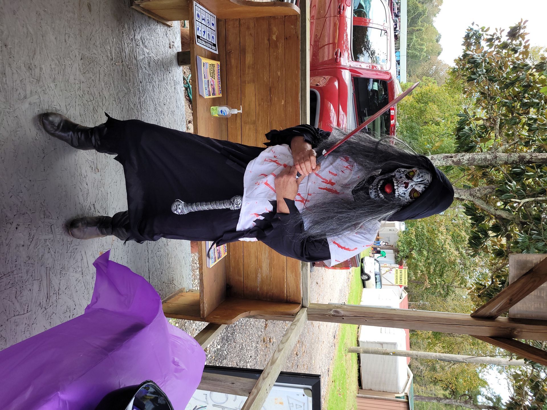 Person in black outfit holding a bloody prop in a rustic outdoor setting with benches and trees