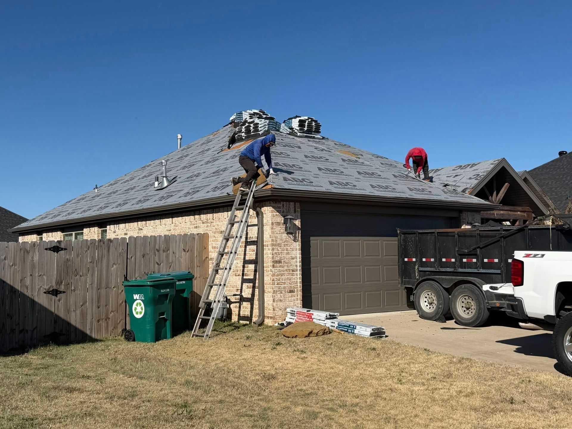 Two workers in blue and red clothing replace shingles on a residential roof next to a trailer and a wooden fence.