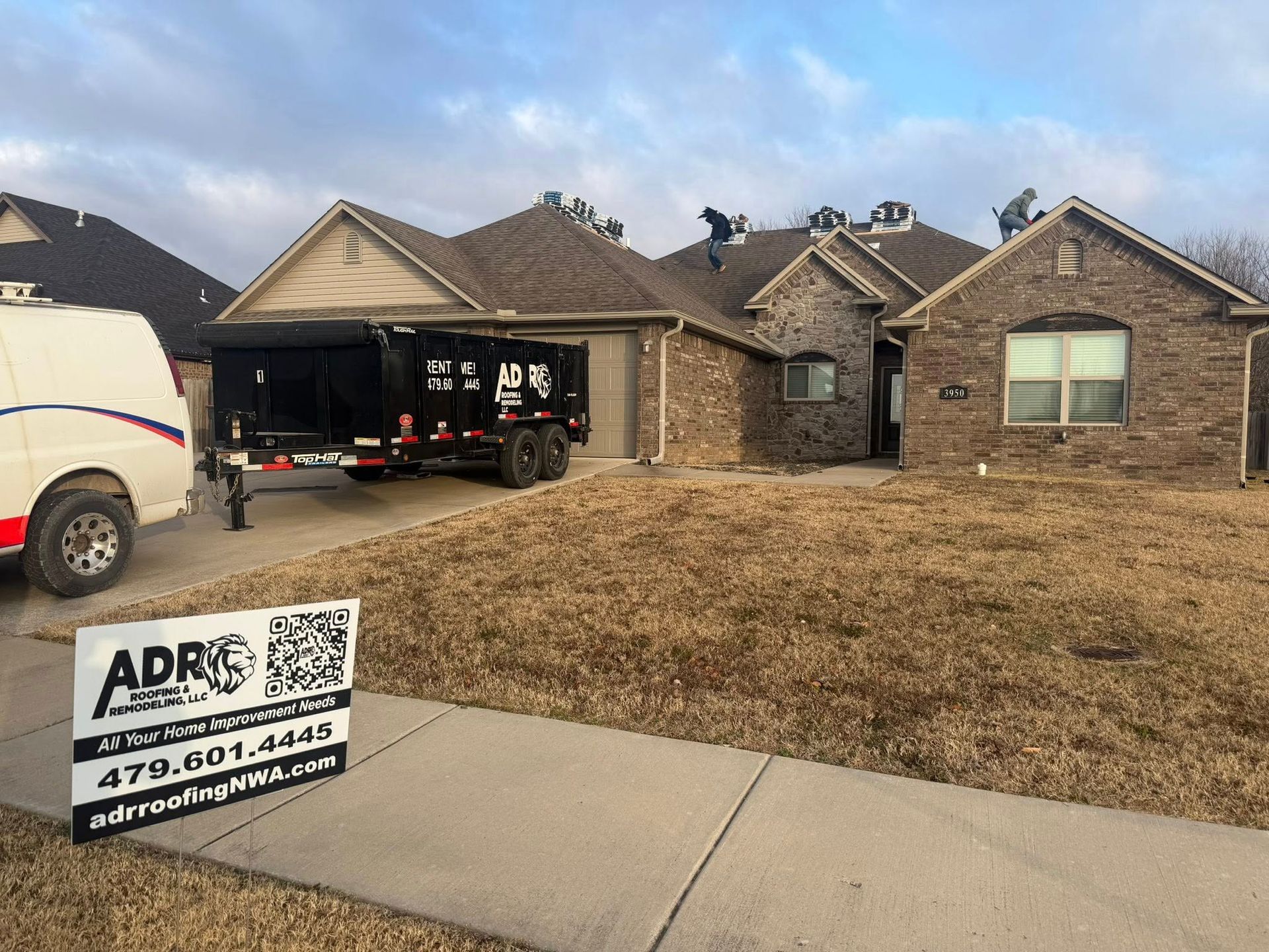 Workers replace a residential roof, with a dumpster trailer and an ADR Roofing sign in the foreground.