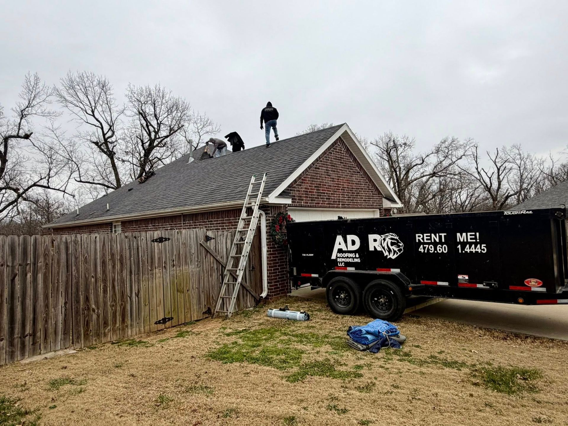 Workers replace the shingles on a residential roof next to a wooden fence and a black dumpster rental trailer.