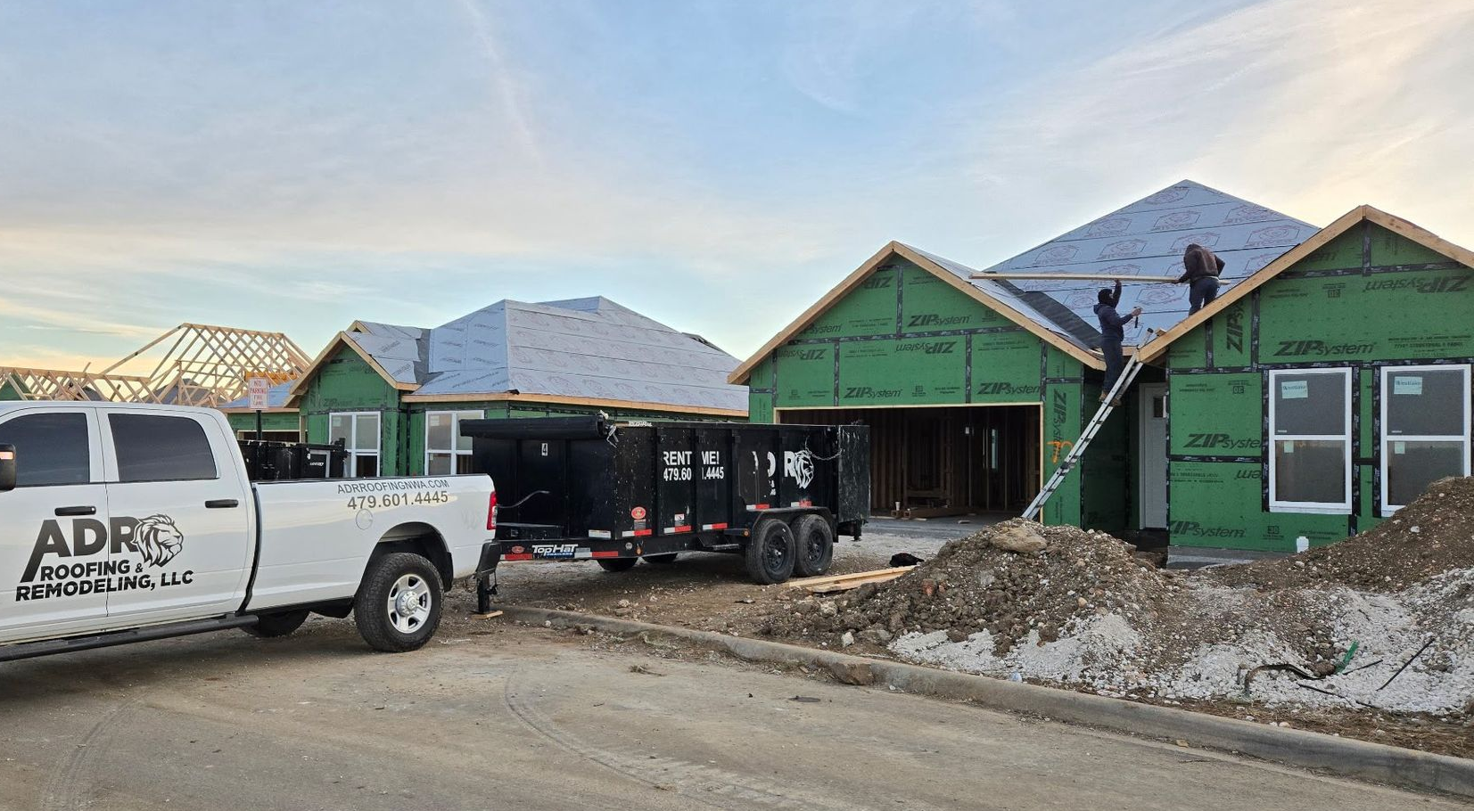 Workers install roofing on new homes with green exterior walls under construction in a suburban development.