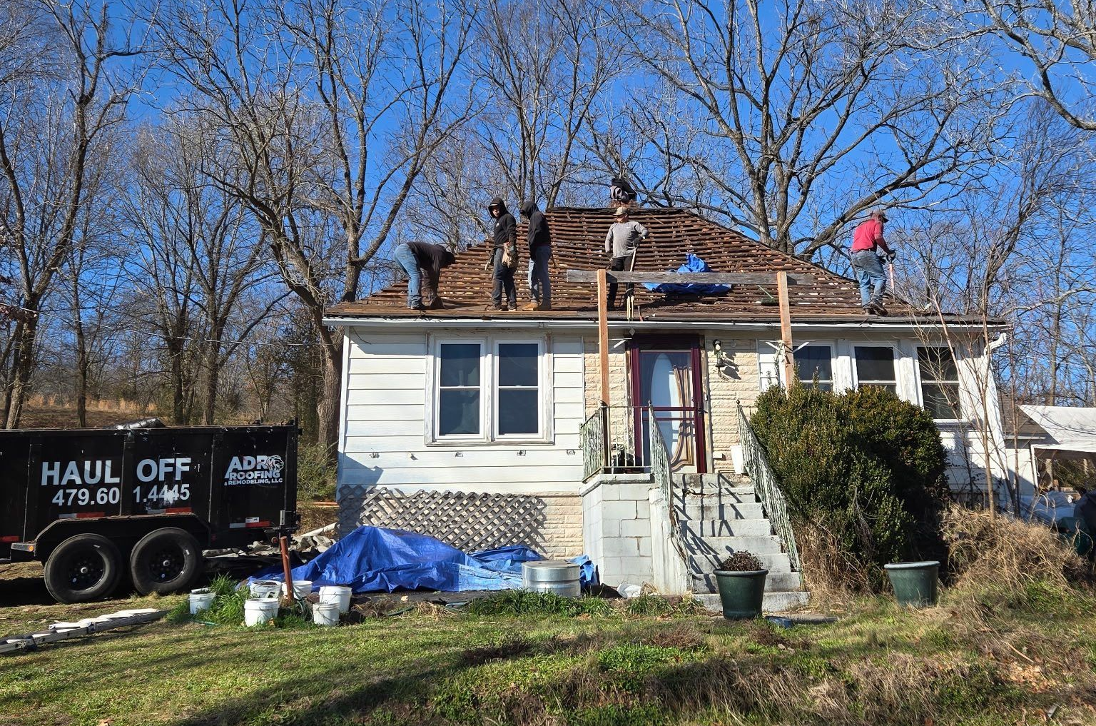 Four people work on stripping shingles from the roof of a small white house, with a debris trailer parked on the left.