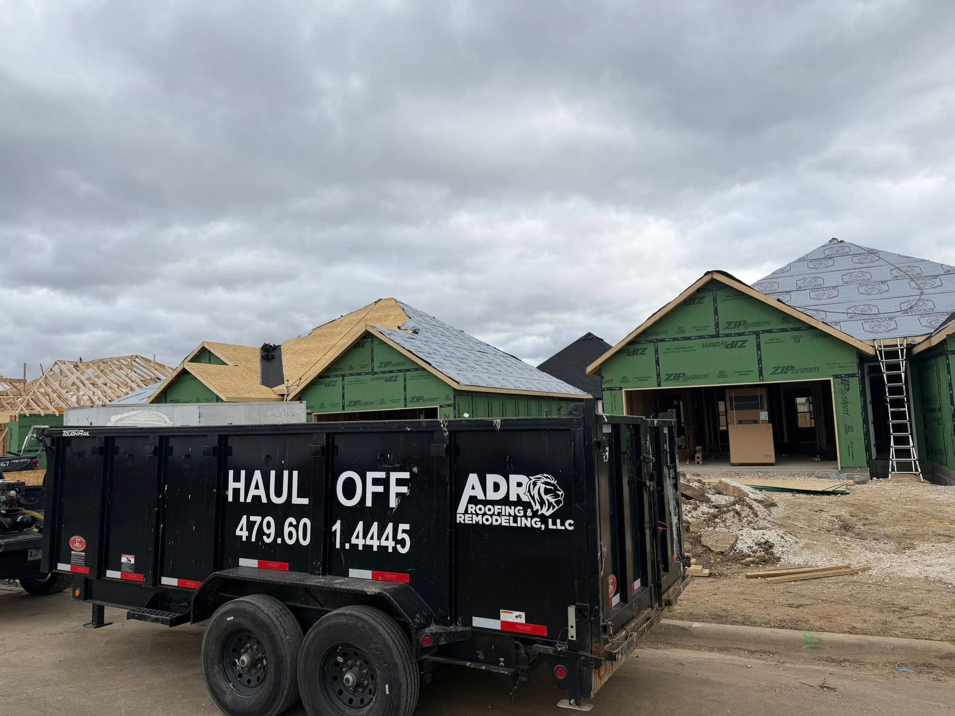 A black ADR Haul Off dumpster sits in front of two unfinished wood-framed houses under a cloudy sky.