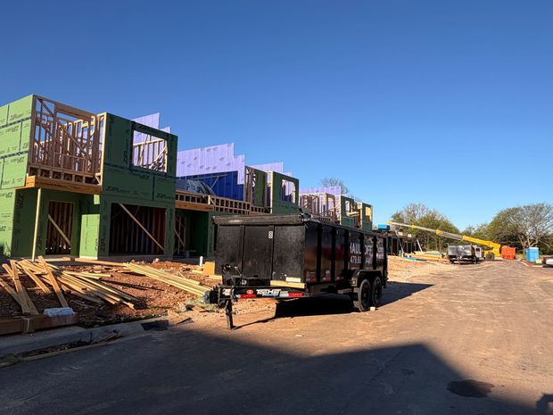 A construction site with several wooden townhomes under construction, a black dump trailer, and a clear blue sky.