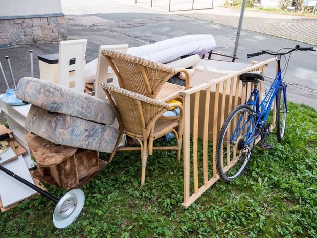 A blue bicycle leans against a pile of discarded furniture, including wicker chairs, cushions, and wooden panels.