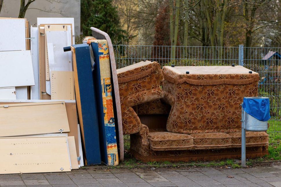 A pile of discarded furniture including a patterned sofa, mattress, and wooden panels left on a sidewalk.