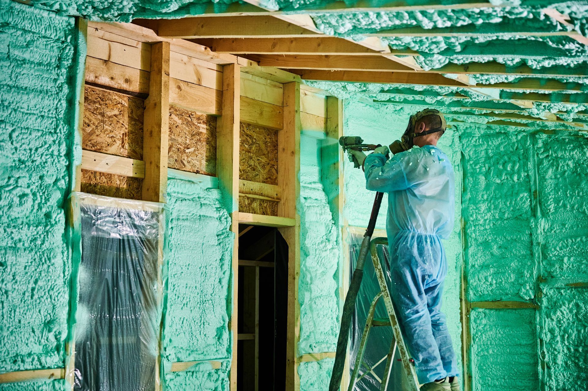 Person spraying green foam insulation inside a building frame, wearing protective gear