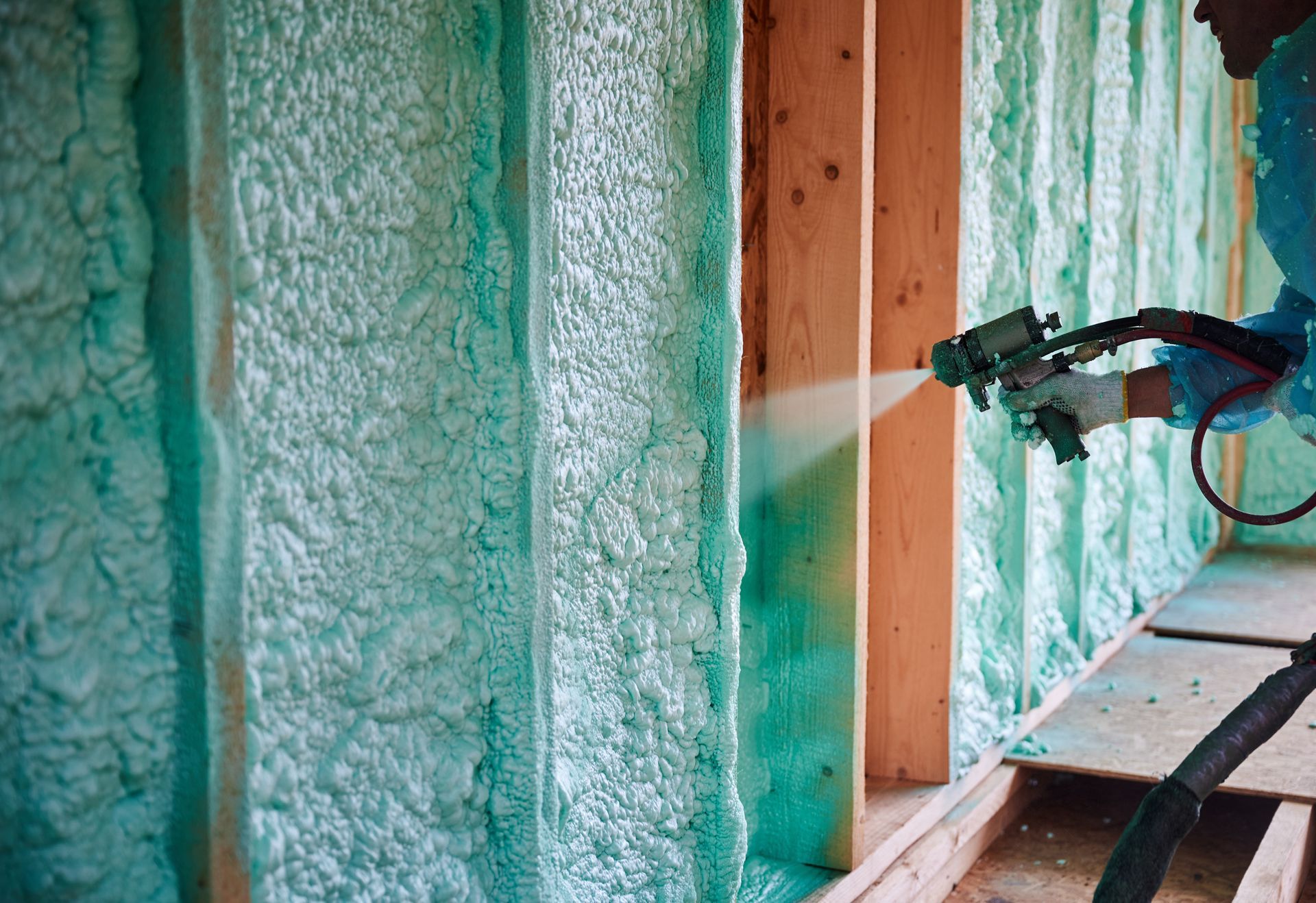 Man sprays green foam insulation between wooden studs of a wall in a construction setting