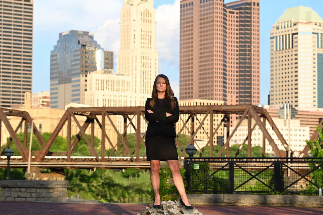 A woman in a black dress is standing on a bridge in front of a city skyline.