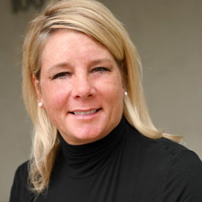 A woman with blonde hair is wearing a black turtleneck and smiling.