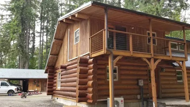 A log cabin with a balcony and a carport in the woods.