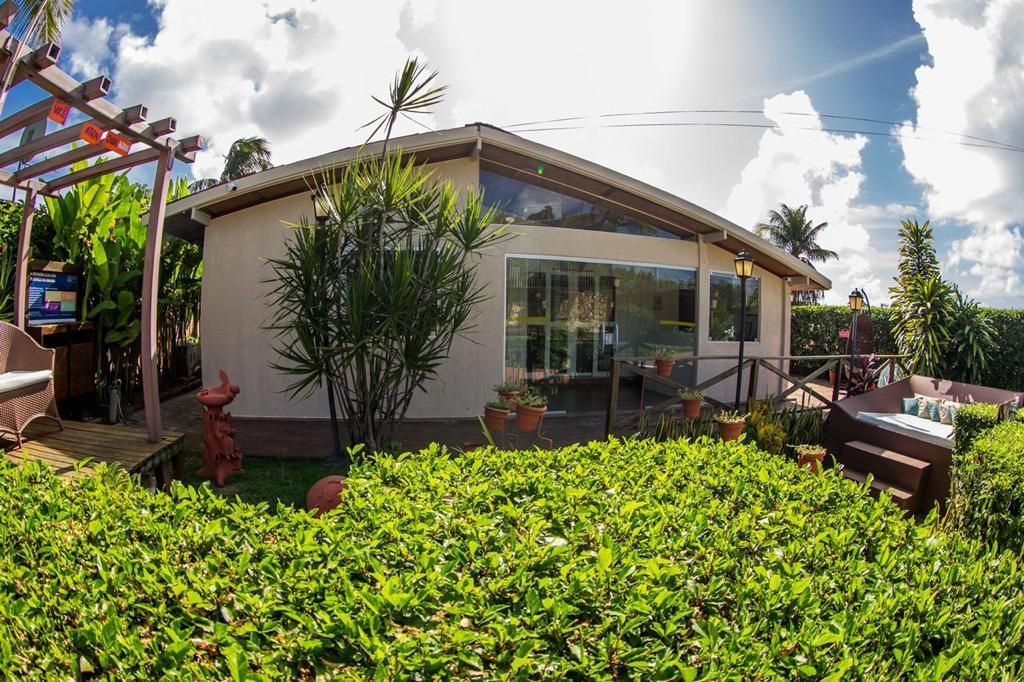 Uma casa com um quintal cheio de vegetação, com grandes janelas e uma pérgola à esquerda.