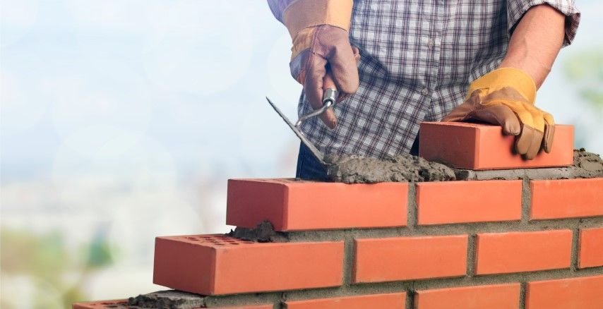 Person laying red bricks with a trowel and mortar on a wall outdoors