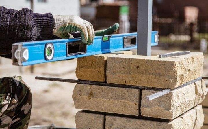 Gloved worker leveling stacked bricks with a blue spirit level at a construction site