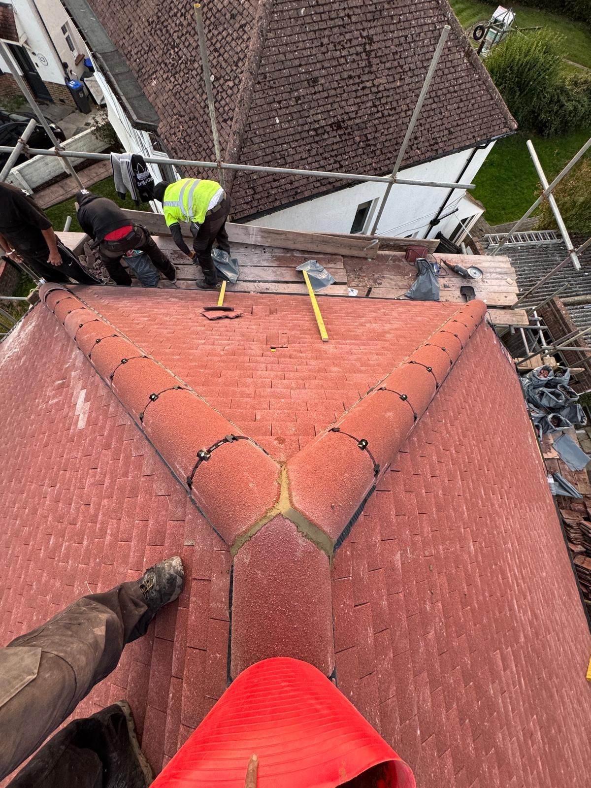 Brick building under construction, roof partially tiled with red and green tiles, scaffolding visible.