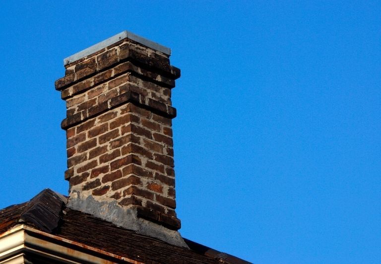 Stone chimney on a gray shingled roof with damaged shingles.