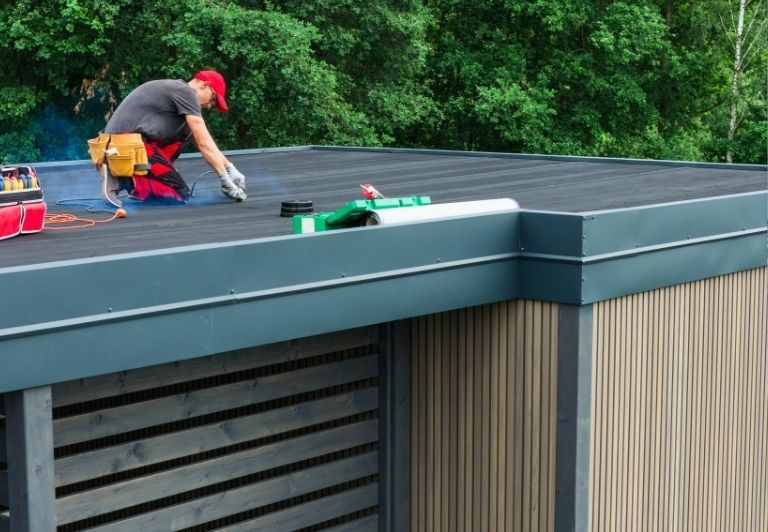 Roofer working on flat black roof with exposed tan base. Tools, rolls, and structure visible.