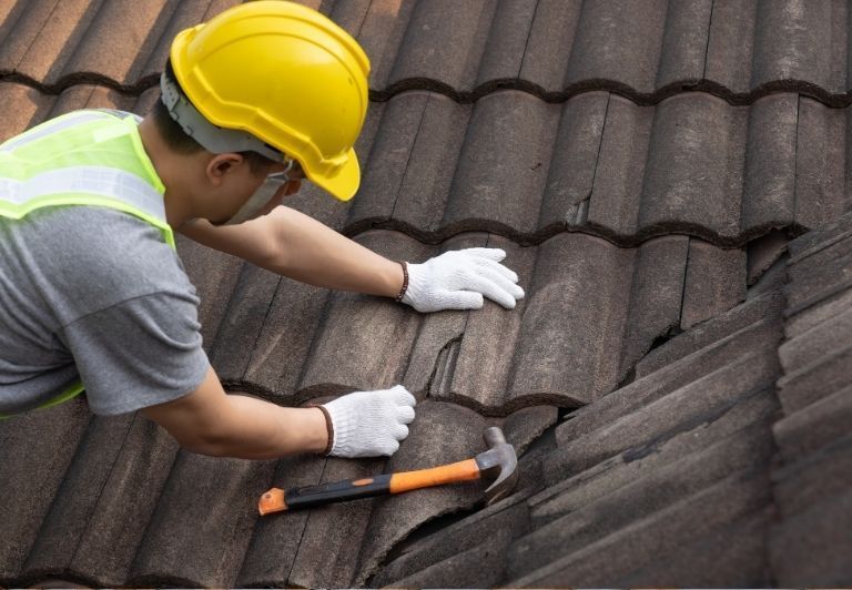 Person wearing gloves patching a cracked brown tile roof with a trowel.