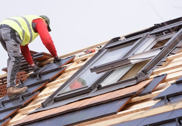 Construction worker, in orange vest and blue helmet, hammering on a rooftop.