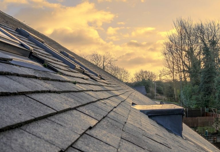 Roofers installing shingles on a house roof. One hammers, the other works in the background. Blue sky overhead.