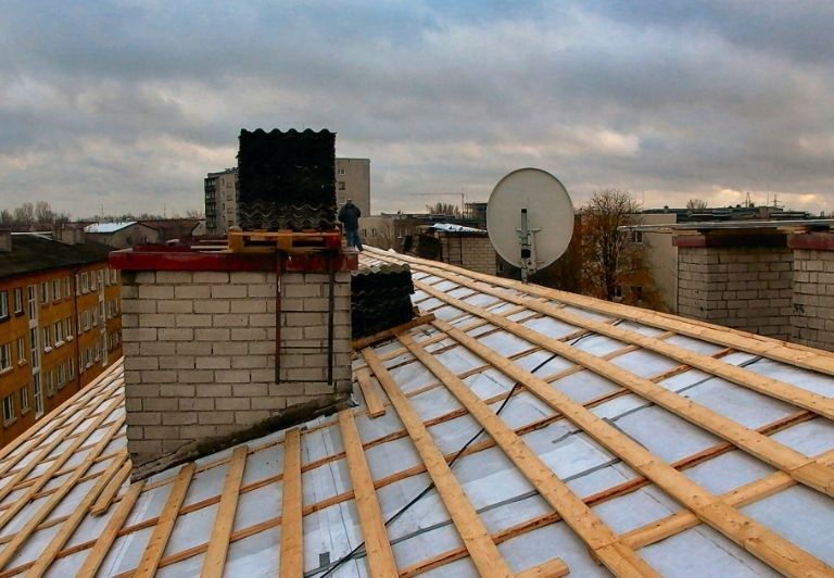 Roofer in hard hat and safety vest repairs roof tiles with hammer.
