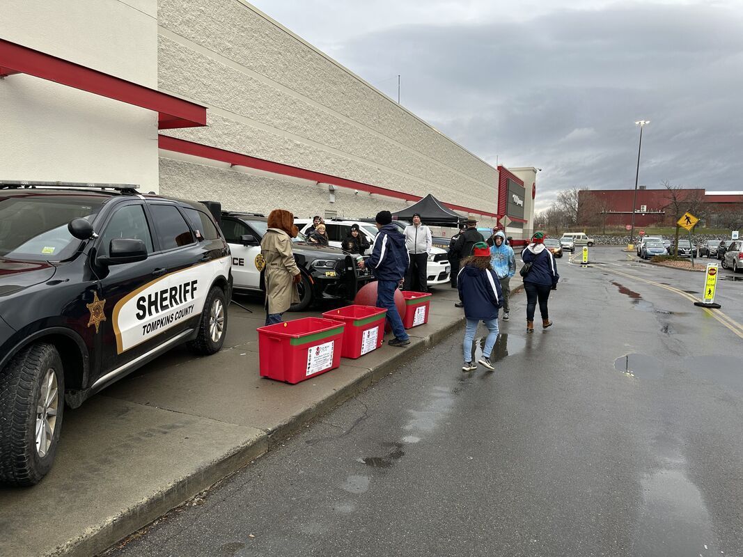 A group of people are standing next to a sheriff 's car in a parking lot near red bins of donated toys.