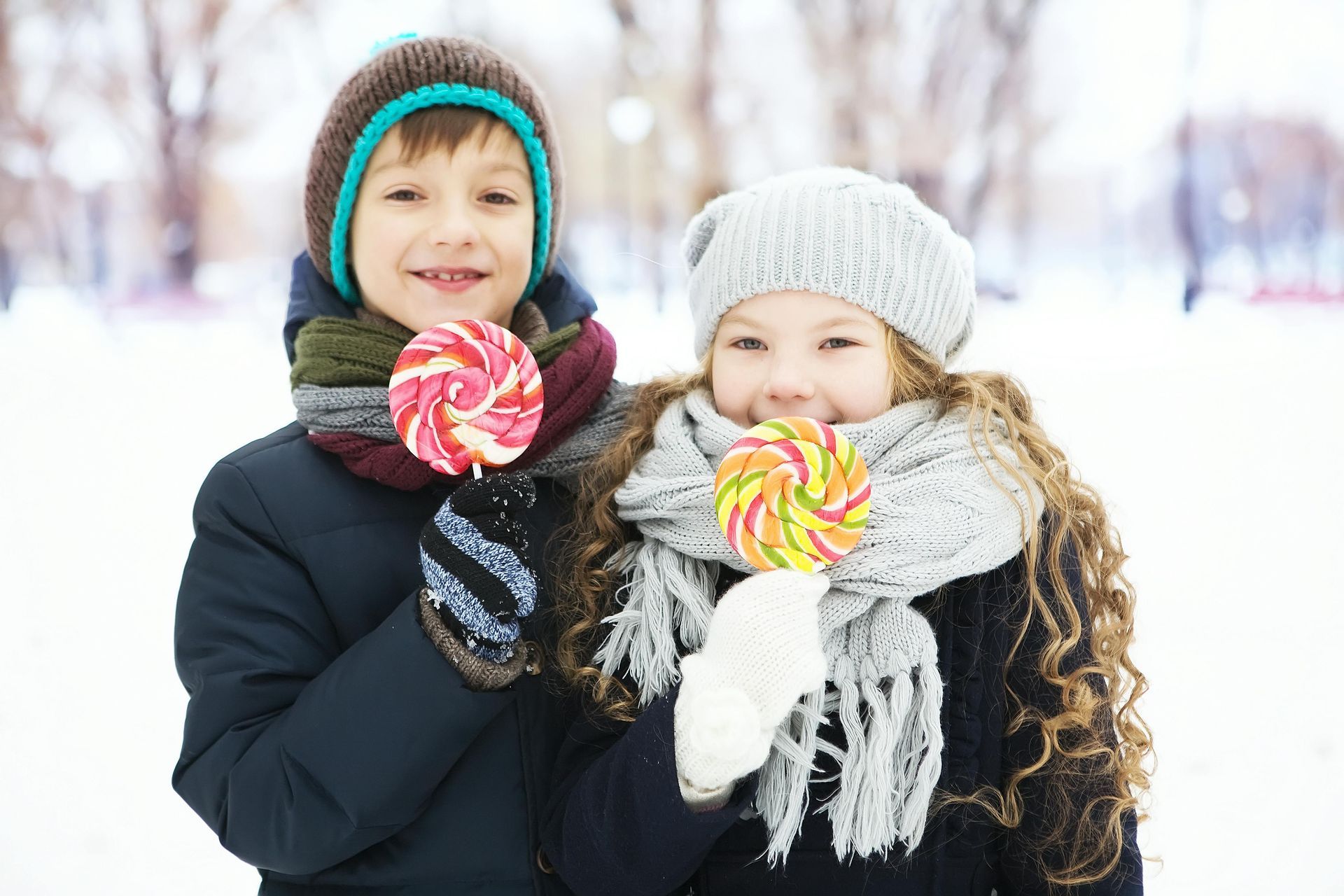 A boy and a girl are eating lollipops in the snow wearing knitted hats, scarves and mittens.