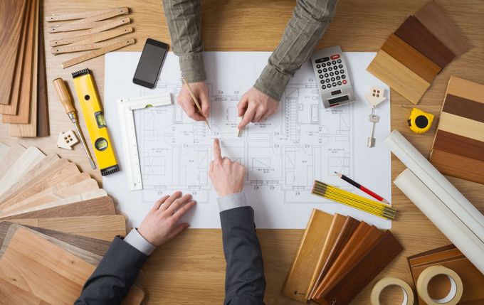 Hands working over architectural plans on a wooden desk with tools and wood samples